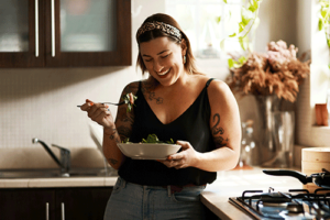A woman is standing in a kitchen and eating from a bowl. She is benefiting from nutritional counseling in San Diego, CA.