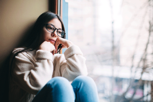 A woman is sitting next to a window and experiencing seasonal affective disorder during the winter months.